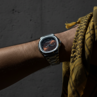 Person wearing a brown arabic dial watch with a stainless steel strap and brown face, draped in a patterned garment against a dark background.
